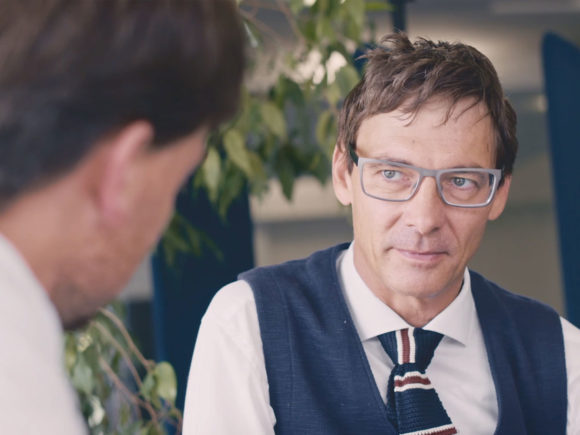 Ein Mann mit Brille und Anzug spricht mit einer anderen Person in einem Innenraum.