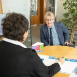 Zwei Personen in einem Büro, eine sitzt am Tisch und die andere steht lächelnd gegenüber.