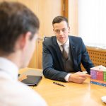 Zwei Männer in Anzügen sitzen an einem Tisch während eines Meetings mit Dokumenten.
