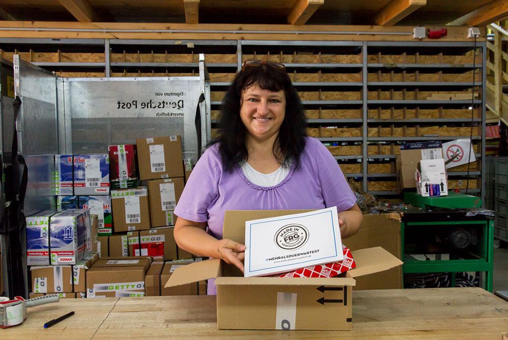 Frau, die ein geöffnetes Paket in einer Lagerhalle mit vielen Kartons hält.