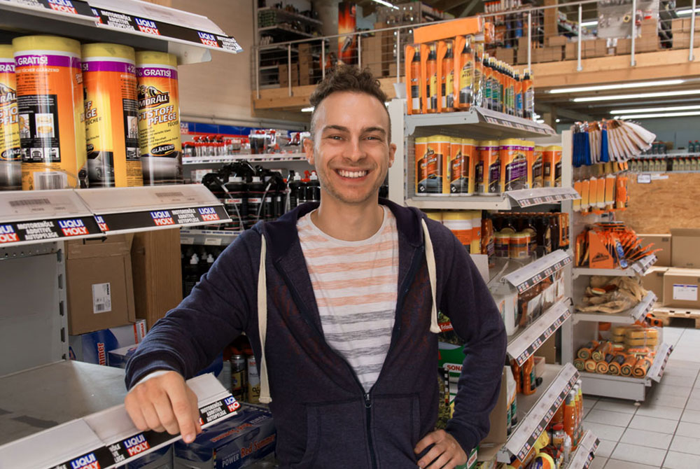 Lächelnder Mann steht in einem Baumarkt mit Regalen voller Produkte im Hintergrund.