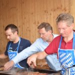 Drei Männer in blauen Schürzen, die an einem Grill Fleisch zubereiten.