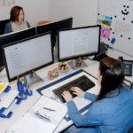 Zwei Personen in einem Büro, eine arbeitet am Computer, die andere sitzt gegenüber.