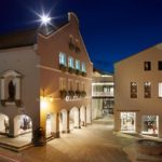 Beleuchtetes Gebäude mit Schaufenstern und Pflanzen an der Fassade in einer städtischen Umgebung bei Nacht.