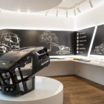 Modell einer Traktorkabine aus schwarzem Kunststoff in einem Ausstellungsraum mit technischen Zeichnungen im Hintergrund.