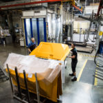 Zwei Arbeiter heben ein gelbes Bauteil in einer Fabrikhalle mit Maschinen im Hintergrund.