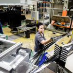 Arbeiter in einer Produktionshalle, der ein Bauteil mit beiden Händen hält und auf eine Maschine blickt.
