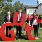 Vier Personen in formeller Kleidung halten große rote Buchstaben, die das Wort 'Gut' bilden.