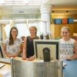 Drei Frauen stehen an einem Empfangstresen mit Computern in einem modernen Büro.