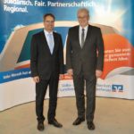 Zwei Männer in formeller Kleidung stehen vor einem Banner mit Text und Logo einer Bank.