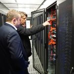 Zwei Personen betrachten Netzwerkgeräte in einem Serverraum mit offenen Schränken und Kabeln.