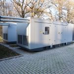 Zwei graue Stromversorgungseinheiten mit Rohren auf der Oberseite, platziert auf einem gepflasterten Boden.