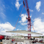 Ein Kran hebt ein großes Betonelement auf einer Baustelle unter einem blauen Himmel.