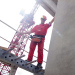 Ein Arbeiter in roter Arbeitskleidung steht auf einem Gerüst und greift nach einem Betonblock.