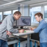 Zwei Männer, die an einem Stehpult arbeiten und einen Computerbildschirm mit einem Fahrzeugmodell betrachten.