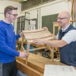Zwei Personen in einer Werkstatt halten einen Holzstuhlrahmen und betrachten ihn.