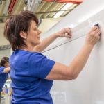 Frau in blauer Kleidung, die Folie an einer weißen Oberfläche in einer Produktionshalle anbringt.