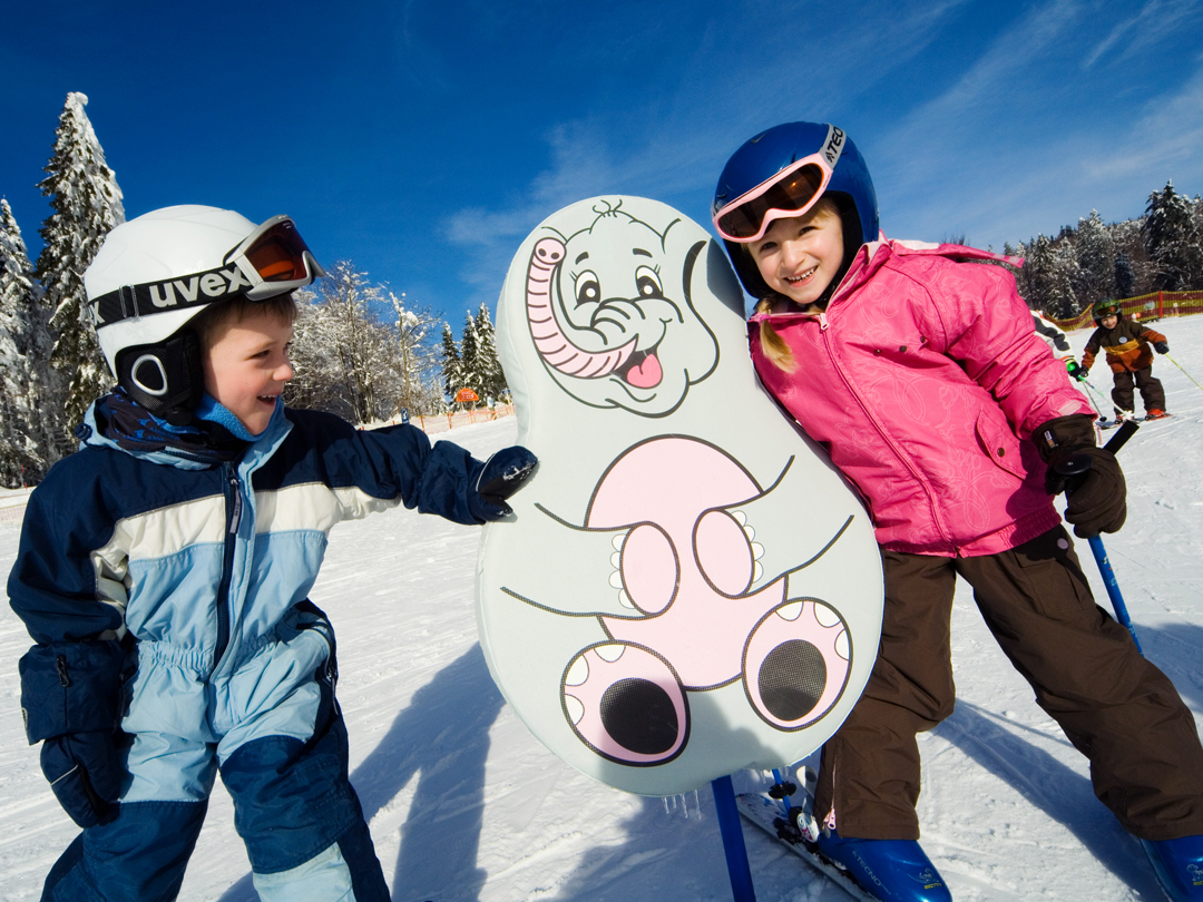 Zwei Kinder in Skiausrüstung stehen neben einem Schild mit einem Elefantenmotiv auf einer Skipiste.