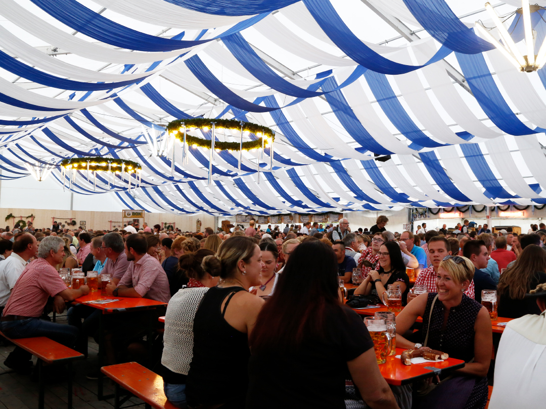 Ein Festzelt mit blauen und weißen Stoffbahnen und vielen Menschen, die an Tischen sitzen und Getränke halten.