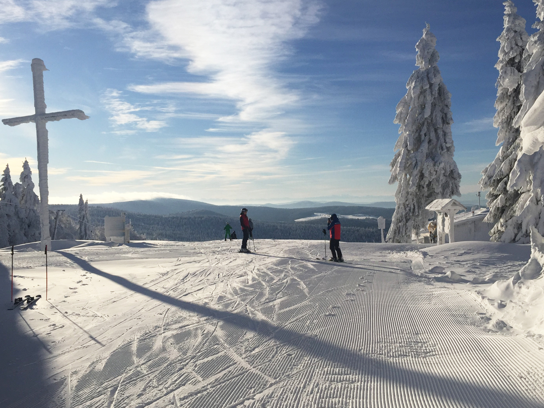 Zwei Skifahrer auf einer verschneiten Piste mit Bäumen und einem Kreuz im Hintergrund.