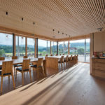 Ein heller Innenraum mit Holztischen und Stühlen, großen Fenstern und Holzdecken.