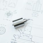 Ein metallisches Bauteil liegt auf technischen Zeichnungen mit Maßangaben und Skizzen.