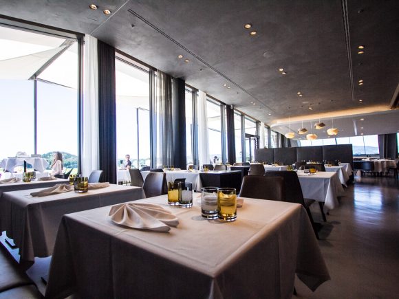Innenansicht eines modernen Restaurants mit Tischen, Stühlen und großen Fenstern.