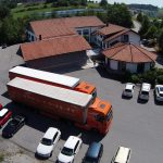 Ein orangefarbener Lkw steht vor einem modernen Firmengebäude mit einem Parkplatz voller Autos.