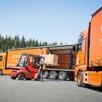 Ein roter Gabelstapler lädt Kartons von einem orangefarbenen Lkw auf einem Parkplatz.