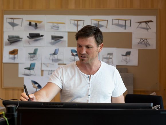 Mann in einem weißen T-Shirt, der an einem Computer arbeitet und Skizzen von Möbeln im Hintergrund hat.