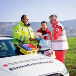 Drei Rettungskräfte des Bayerischen Roten Kreuzes stehen neben einem Einsatzfahrzeug und betrachten eine Karte.