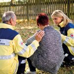 Zwei Rettungskräfte des Roten Kreuzes helfen einer Person, die auf dem Boden sitzt und Unterstützung benötigt.