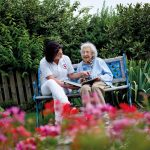 Eine Pflegekraft sitzt neben einer älteren Dame auf einer Bank im Garten und zeigt ihr ein Tablet.