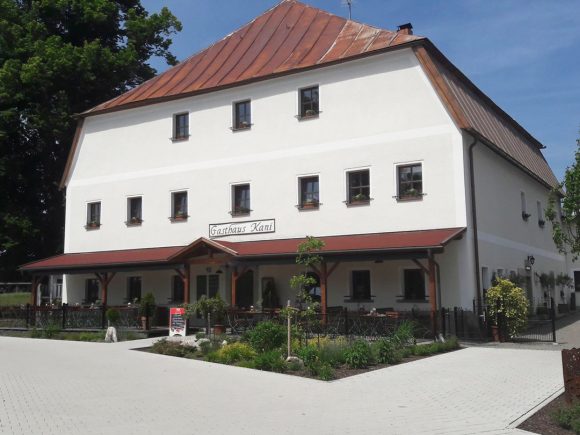 Gasthaus Kani mit weißer Fassade und rotem Dach in einer ländlichen Umgebung.