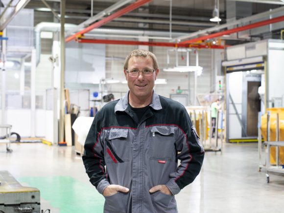 Mann in grauer Arbeitskleidung mit Brille steht in einer Fabrikhalle und lächelt.