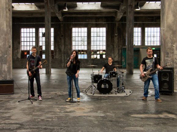 Eine Band mit vier Mitgliedern, die in einem Industriegebäude auftritt, mit Instrumenten wie Gitarre, Bass und Schlagzeug.