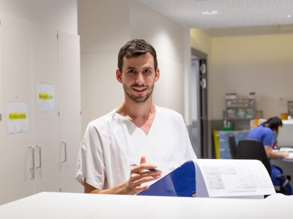 Ein Mann in medizinischer Kleidung hält Dokumente und lächelt am Empfang eines Gesundheitszentrums.