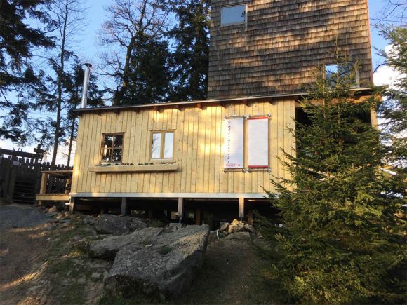 Holzgebäude mit Fenstern und Schindeldach in einer bewaldeten Umgebung.