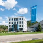 Moderne Bürogebäude von Knauf mit blauer Flagge, umgeben von Grünflächen und einem klaren Himmel, ideal für Unternehmenspräsentationen.