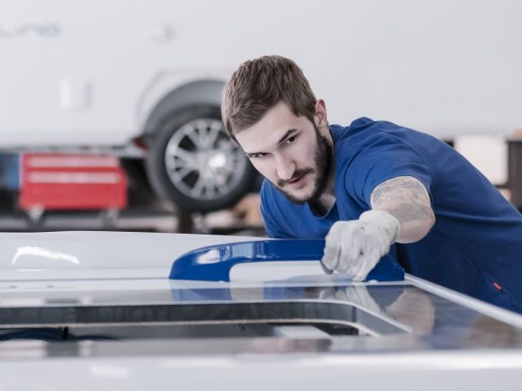 Ein Mitarbeiter mit Handschuhen bearbeitet das Dach eines Fahrzeugs in einer Werkstatt.