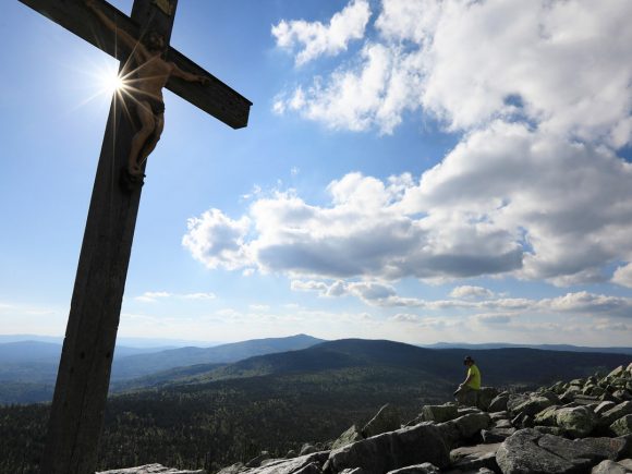 Holzkreuz mit einer Figur am Lusen, umgeben von einer Berglandschaft unter einem blauen Himmel.