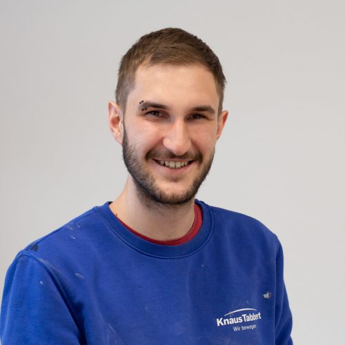 Mann mit kurzem Bart und lächelndem Gesicht, trägt ein blaues Sweatshirt mit Logo.