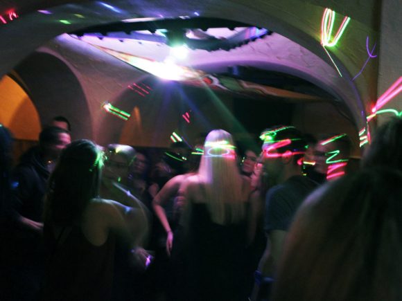 Menschen tanzen in einem Musikclub mit bunten Lichtern und einer dynamischen Atmosphäre.