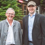Zwei Männer stehen nebeneinander, einer trägt eine traditionelle Tracht, der andere einen Anzug mit Hemd und Kappe.