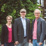 Drei Personen stehen nebeneinander im Freien, zwei Männer und eine Frau, alle lächeln.