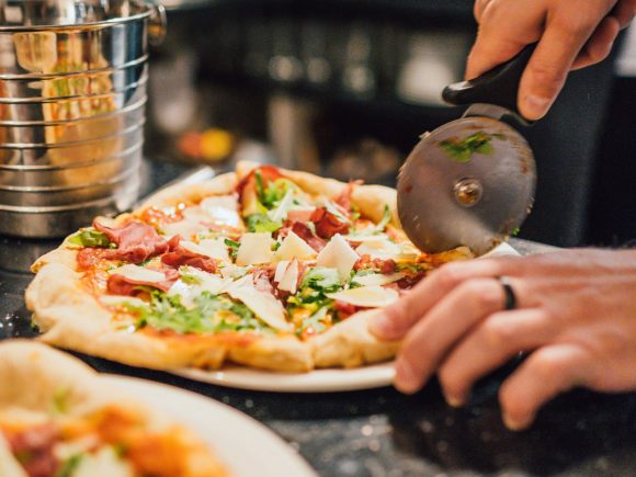 Eine Hand schneidet eine Pizza mit Schinken, Käse und frischem Koriander mit einem Pizzaschneider.