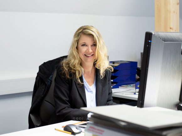 Frau mit blonden Haaren sitzt an einem Schreibtisch und arbeitet am Computer.