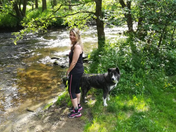 Frau steht neben einem Hund am Ufer eines Flusses in einer grünen Umgebung.
