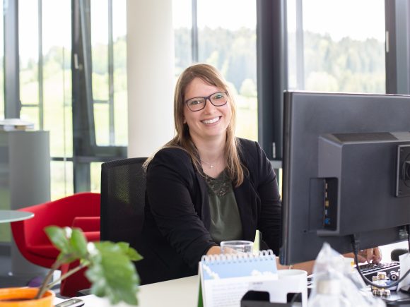 Eine Frau mit Brille sitzt an einem Schreibtisch und lächelt in die Kamera, während sie an einem Computer arbeitet.