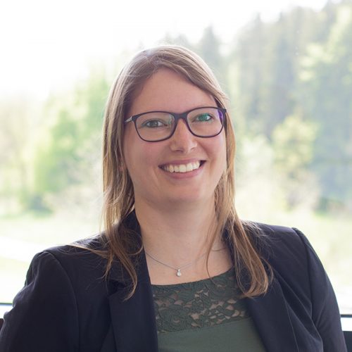 Frau mit langen Haaren und Brille, die in die Kamera lächelt, trägt einen Blazer.